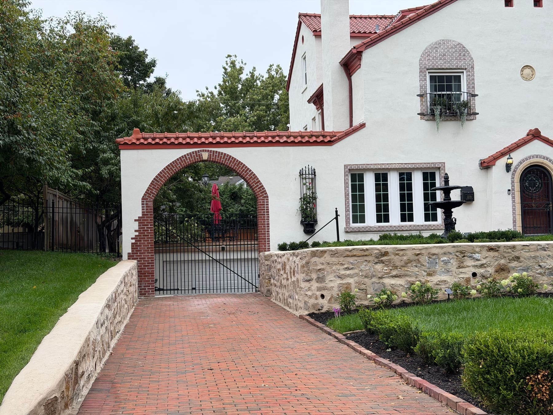 Red brick driveway leading to a brick-lined stucco arch with an iron gate and stone retaining walls